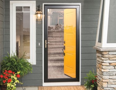 A glass storm door is in front of a yellow front door on a house with gray-blue siding