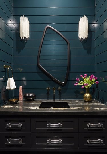 Hollywood regency powder bathroom with glossy teal walls, black marble vanity, and brass, crystal and acrylic details