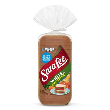 A loaf of white bread in clear packaging. The Sara Lee logo is across the wrapping, with a green label below it that reads "white bread with veggies". There is also a sandwich on a plate, and a carrot, tomato and a squash.