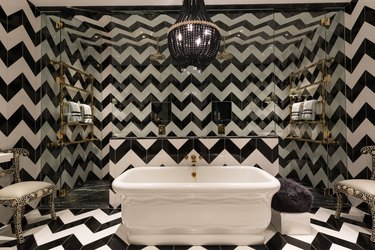 Hollywood Regency bathroom with black and white floor-to-ceiling chevron tile, white freestanding tub and oversize black chandelier.