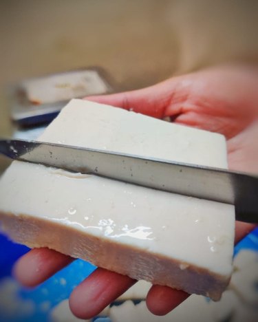A block of tofu in a hand with a knife slicing through it.