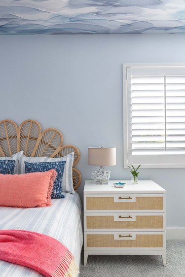 Bedroom with rattan headboard, blue and white watercolor ceiling, white window shutters and bedding in blue, coral and white