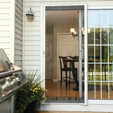 A retractable screen door on the back of a home with white siding; a stainless steel grill is on the porch