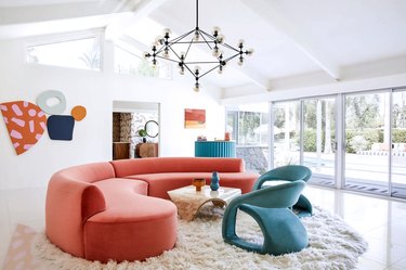 A white room with a curved red couch, curved blue chairs, a triangular chandelier, and colorful art on the walls.
