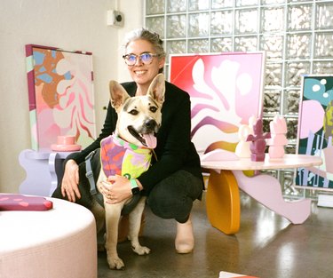 Alex Proba wearing all black and glasses and squatting next to a dog in her studio full of colorful paintings and shape tables.