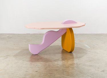 A pink, lavender, and orange shaped table on a beige floor in front of a white wall.
