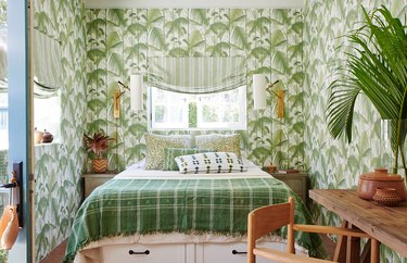 A green and white maximalist bedroom with palm tree wallpaper, striped window treatments, and mixed pattern bedding.