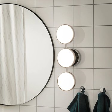 round mirror on white tiles with wall lamp nearby with three bulbs