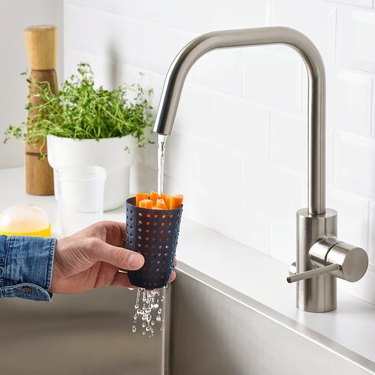 person holding container with vegetables under faucet with water