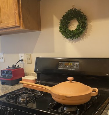 A light pink Always Pan rests on a black gas stove top. There is a small boxwood wreath on the wall above the stove and a blender to the side.