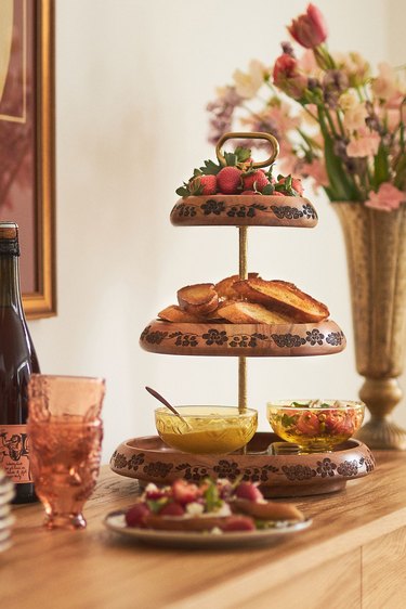 A hand carved three tiered dessert tray rests on a wooden table next to a bouquet of flowers, a bottle of wine, two pink drinking glasses and a plate of strawberry toasts. The top tier has strawberries in it, the second tier has toast and the third tier and a bowl of yogurt and a bowl of strawberries.