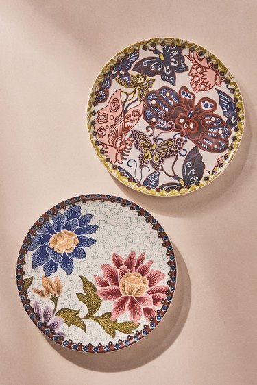 Two dessert plates, the plate in the bottom left corner is light blue with a dark blue flower and dark pink flower, while the plate in the upper right corner is a mix of greens, blues, reds and pink butterflies.