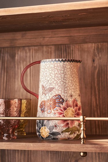 A large white pitcher adorned with striking florals and garden imagery rests on a wooden shelf next to a pink  and yellow glass.