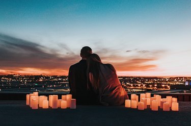 A woman leaning on a man's shoulder facing away from the camera surrounded by candles while watching a sunset over a city.