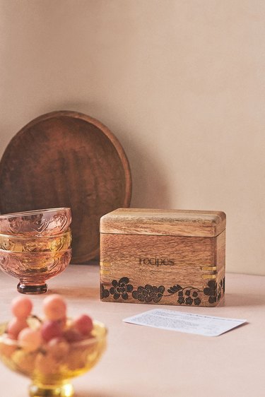 A cedar recipe box rests on a counter next to amber colored stacked glasses, a bowl of red grapes and a wooden bowl that's leaning against the wall.