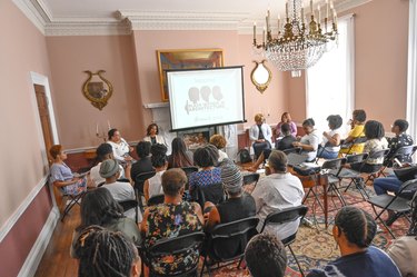A wide shot of a conference room with light pink walls. A crystal chandelier hangs in the middle of the ceiling. A group of Black women are sitting in chairs arranged in front of a projector. The image on the projector shows a logo of three Black women. The text reads: Welcome, Black Women in Architecture, Brunch 2019.