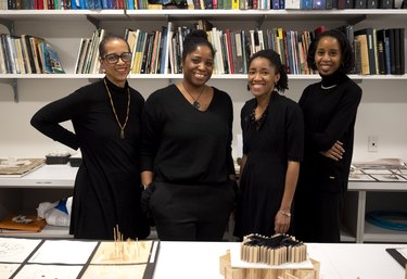 A portrait of the Riding the Vortex collective: four Black women stand in front of an industrial bookshelf filled with books. The four women are smiling and wear various black outfits. In front of them is a table filled with architectural models and designs.
