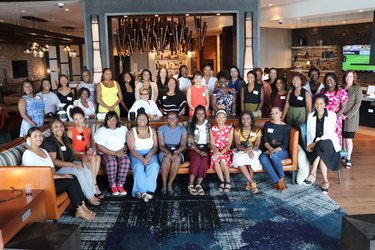 A wide shot of a group of Black women gathered in a hotel lobby. They are sitting on a striped L-shaped couch that's in front of a round blue rug. They are attendees of the D.C. brunch.
