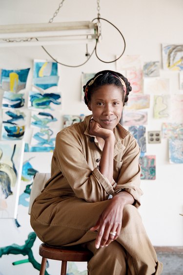 Artist K'era Morgan, a Black woman with short, pulled-back black hair, in a light brown jumpsuit. She is in a studio, in front of white wall covered in her work.