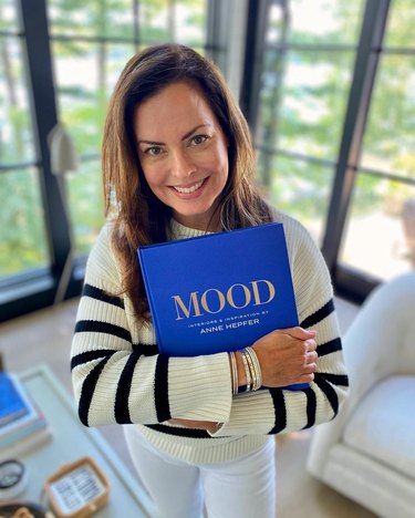 Anne Hepfer in a cream and black striped sweater holding her book "Mood: Interiors & Inspiration"