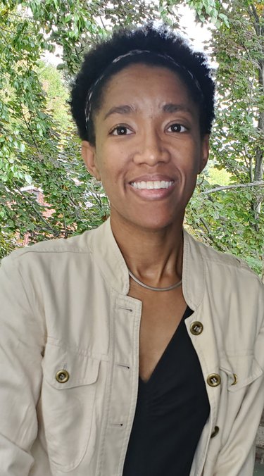 A portrait of Katherine Williams, a Black woman with light brown skin and a short Black afro. She is smiling in front of trees on a sunny day. She is wearing a beige shirt that is open, showing a black T-shirt underneath.