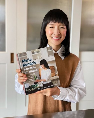 Marie Kondo in a white and brown shirt holding up a book titled "Marie Kondo's Kurashi at Home: How to Organize Your Space and Achieve Your Ideal Life (The Life Changing Magic of Tidying Up)"