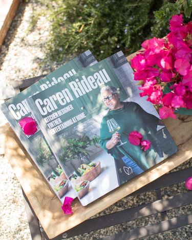 Caren Rideau's book "Caren Rideau: Kitchen Designer, Vintner, Entertaining at Home" on a wooden board next to pink flowers and green shrubs