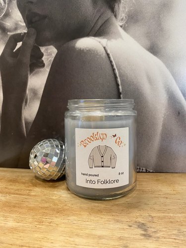 A small Brooklyn + Co candle rests on a wooden surface next to aa mini disco ball and in front of a Taylor Swift Folklore vinyl. The candle features a label with Taylor Swift's white cardigan on the front, and the candle is labeled "Into Folklore"