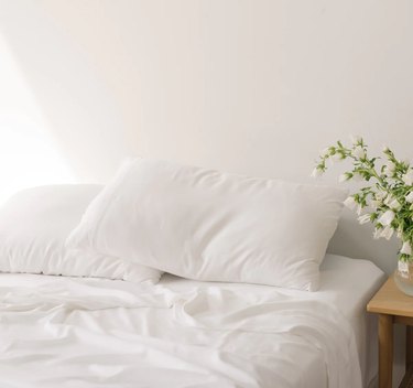 white sheets on an unkempt bed; nightstand with white flowers