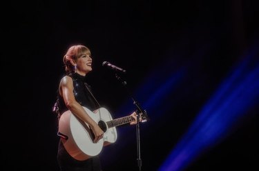 Taylor Swift stands smiling on stage with a guitar over her shoulder in front of a microphone