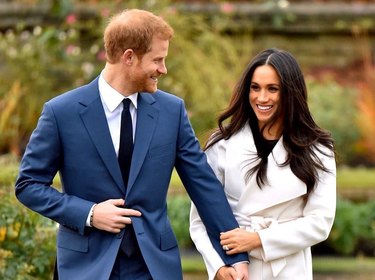Prince Harry in a navy blue suit smiling at Meghan Markle in a cream-colored pea coat while they hold hands.