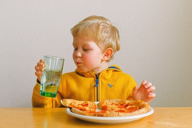 Child with blond hair drinking water and eating pizza at a table