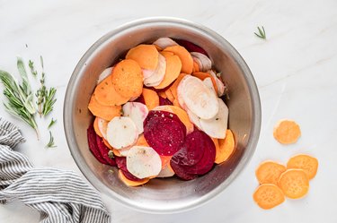 The sliced sweet potatoes, rutabagas, and beets in a metal bowl on a white marble countertop.