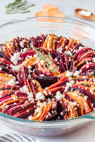 A root vegetable and goat cheese bake in a glass pie pan featuring red, orange, and dark pink root vegetables sprinkled with goat cheese.