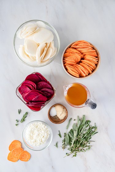 The sweet potato slices, rutabaga slices, beet slices, vegetable broth, herbs, and spices in small bowls on a white marble countertop.