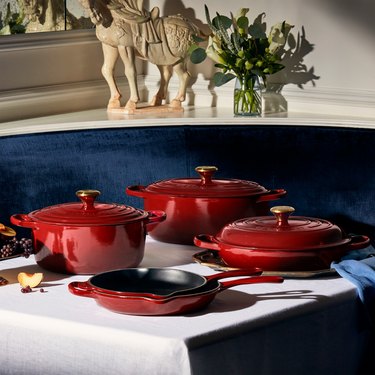 Four dark red Le Creuset pots and pans on a dinner table with a white tablecloth and a small tabletop horse statue behind it.