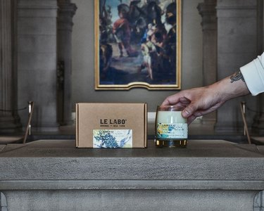 Hand holding the Le Labo and The Met candle next to a cardboard Le Labo box in front of an old painting.