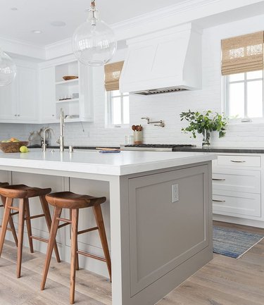 A bright white kitchen with greige island, warm wood barstools and blue throw rug.