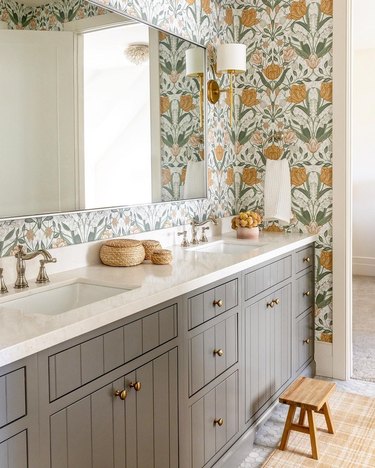 A bathroom with mustard and green floral wallpaper, warm gray cabinets, and wood accents.
