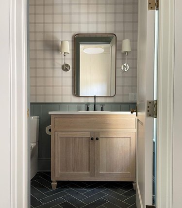 A modern traditional bathroom with plaid wallpaper, sea green wainscoting, and slate herringbone tile.