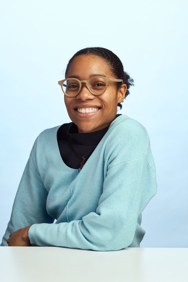 A photo of Tiarra Bell, a Black person with black hair pulled back wearing light brown glasses and a light blue sweater over a black T-shirt.