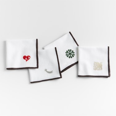 Four white napkins with black edges embroidered at the corner with one of Lucia Eames' favorite designs: her captivating Seeing with the Heart emblem in red, an awe-inspiring silver comet, a stylized green sunburst, and her signature dove stitched in cream.