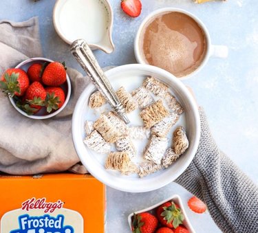 Hand with a gray sleeve holding a white bowl of Frosted Mini Wheats with milk over a light blue countertop with two bowls of strawberries, a cup of milk, and a mug of coffee.