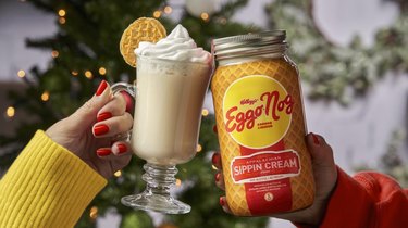 A glass and a jar of Eggo Nog toast in front of a lit Christmas tree: The glass on the left is a tall, clear mug of eggnog, topped with whipped cream and garnished with a mini Eggo waffle, held by a medium-skin toned hand with red nails and a yellow sweater, while the Eggo Nog jar is held by a medium-skin toned hand in a red shirt. The Eggo Nog jar is wrapped with a waffle printed label, a yellow circle with red text reading Kellogg's Eggo Nog, with white text reading "Sippin' Cream" below, with a red background.