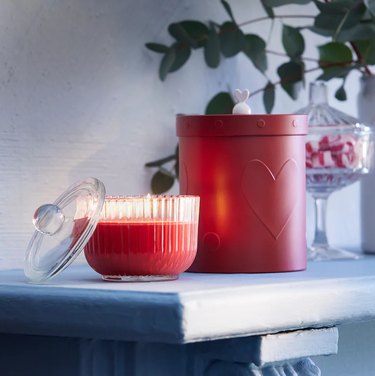 A red candle in a glass fluted jar with a lid on a white mantel next to a red heart canister.