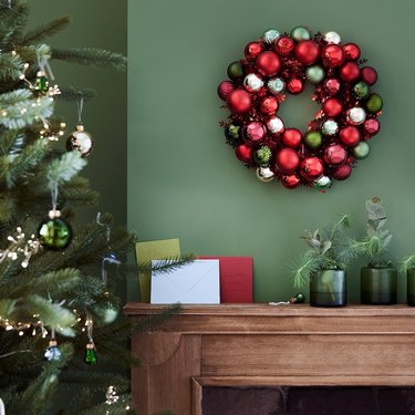 A holiday wreath made up of silver, green, red, and pink ornaments on a green wall next to the Christmas tree.