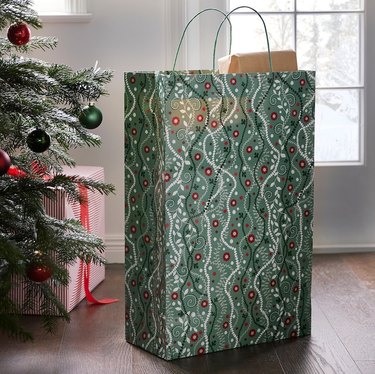 A tall gift bag in a red, green, and white floral holiday pattern next to a Christmas tree on a dark wood floor.