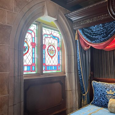 A stained glass window featuring a crown in the Cinderella Castle Suite, next to a canopy bed in gold, blue, and red linens.