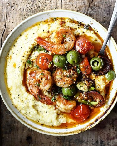 A white bowl filled with cheese polenta, chorizo, shrimp, green olives, and cherry tomatoes.
