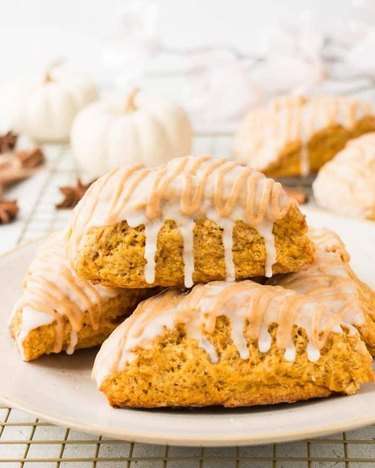 A stack of triangle pumpkin scones drizzled with white icing on a beige plate.